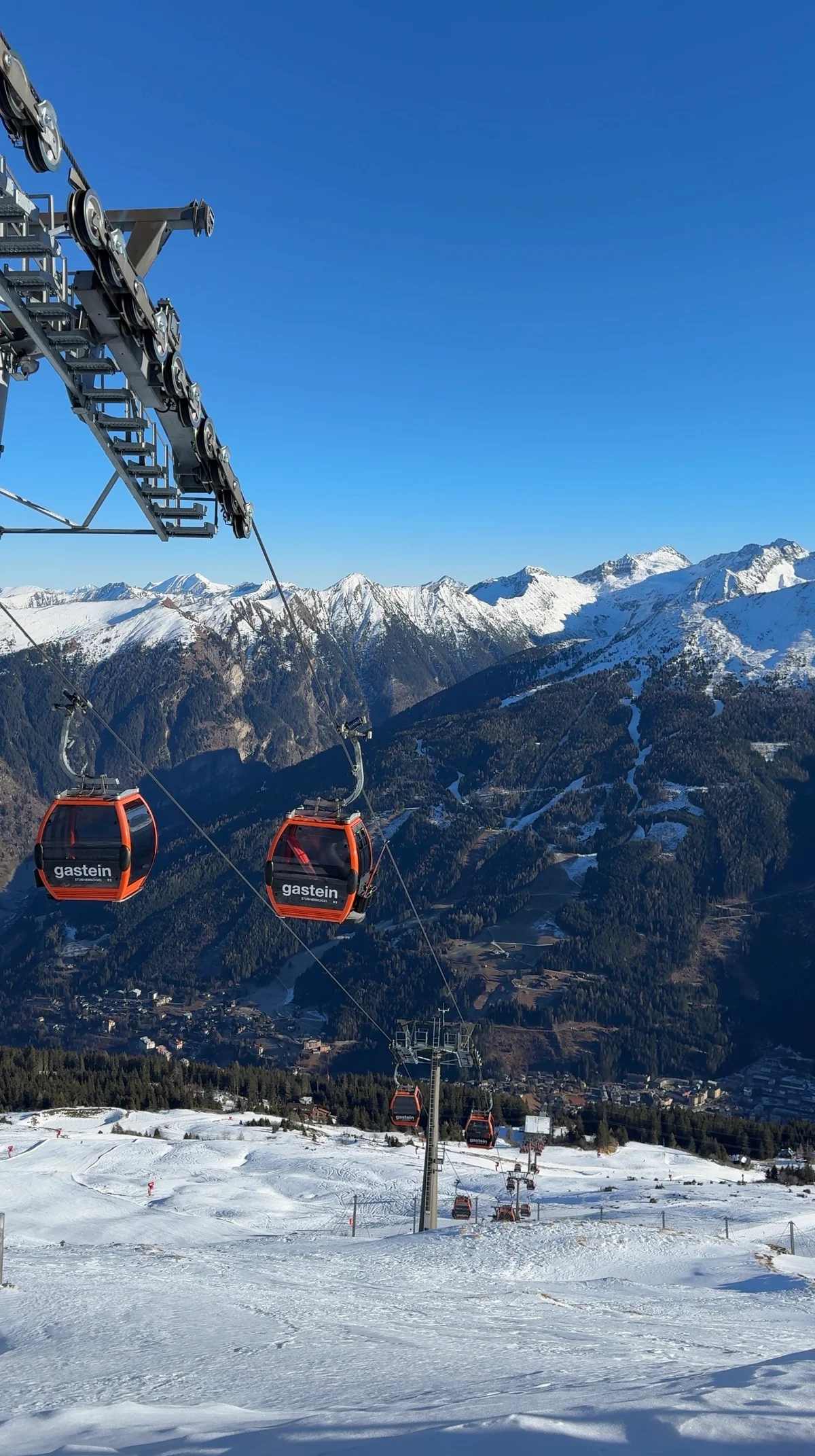 Red Gastein gondolas above a snowy slope, with a valley and mountain ridges in the background.