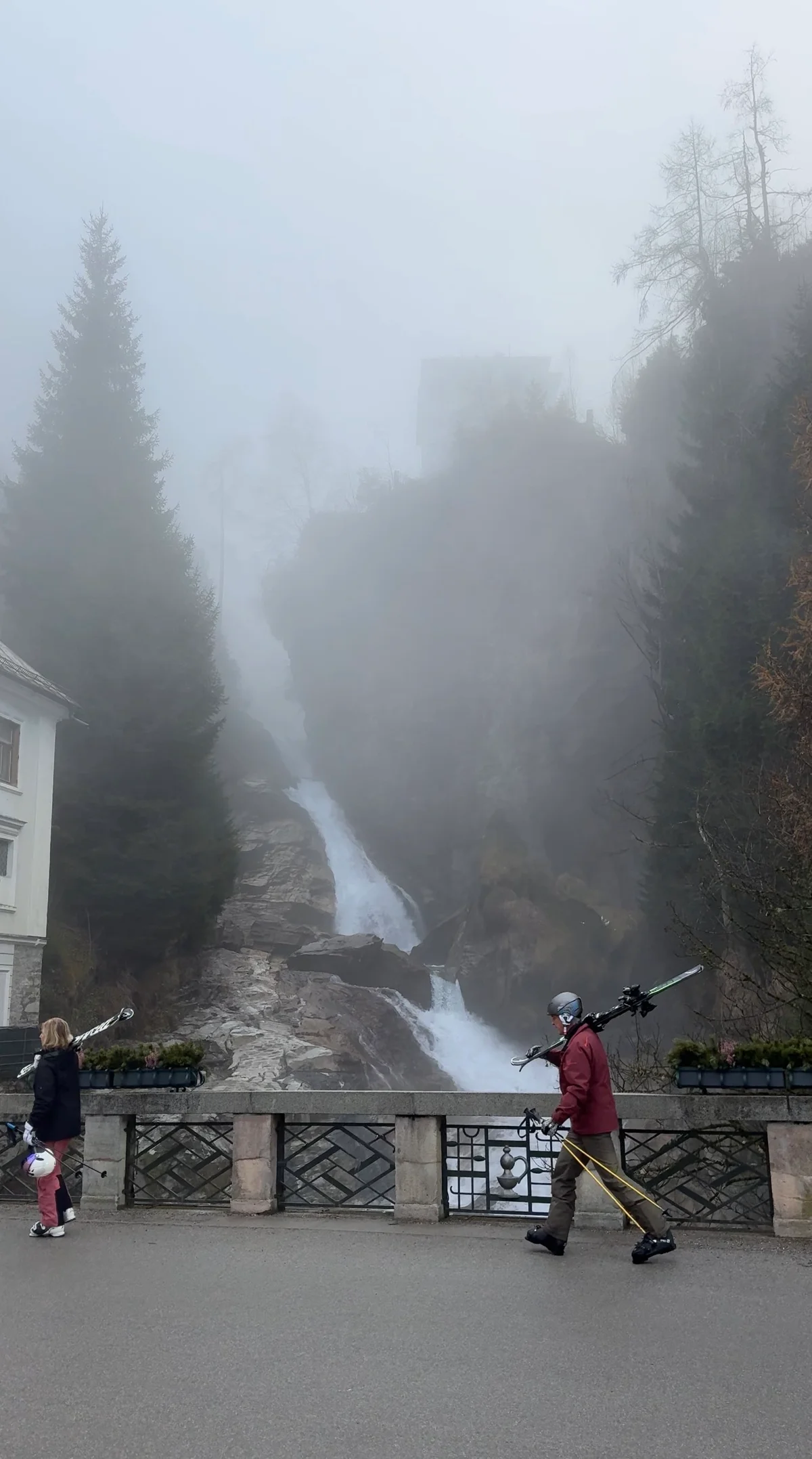 Bad Gastein’s waterfall in mist behind a bridge, with people carrying skis in the foreground.