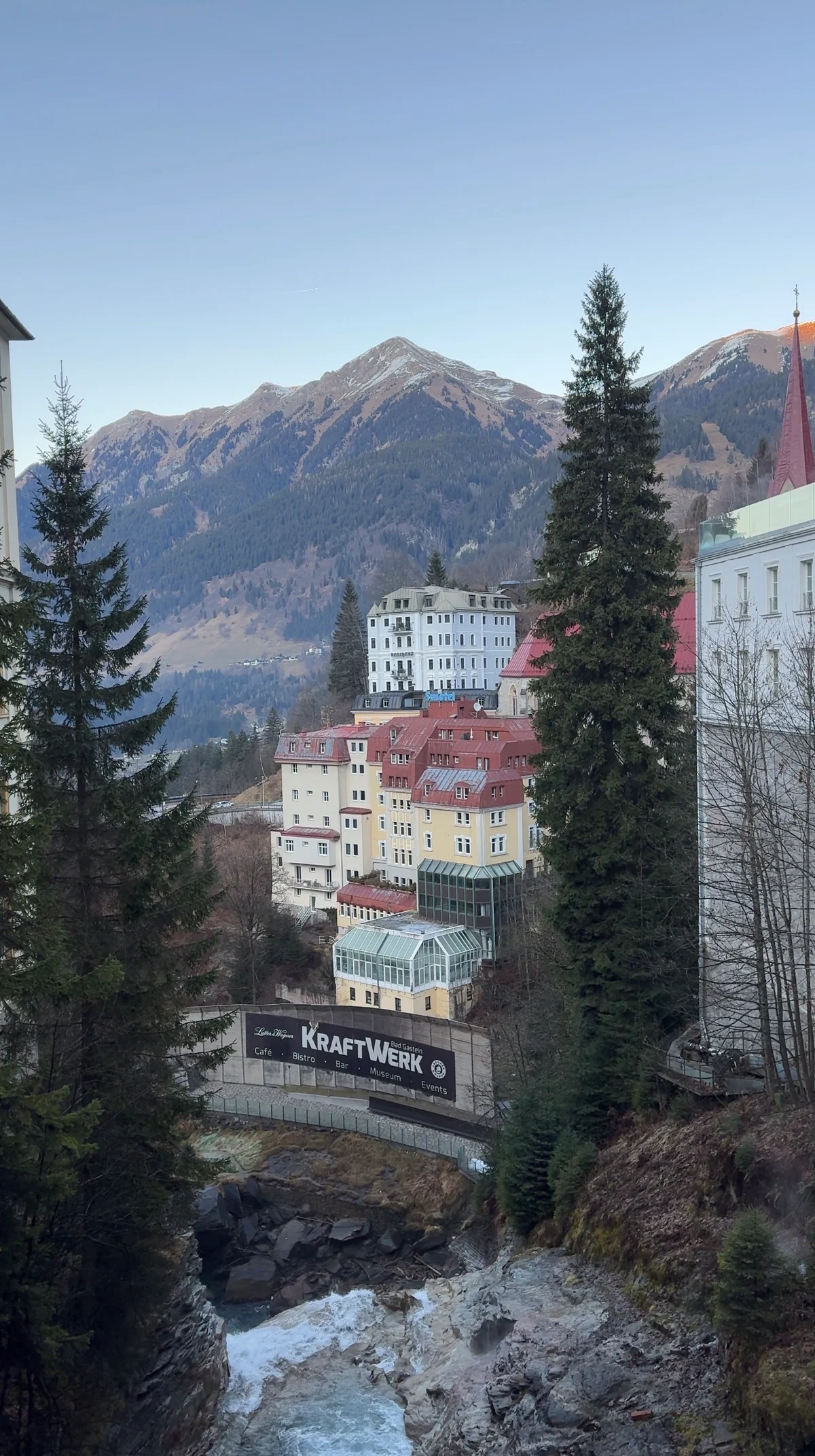 Cascading water in a rocky gorge below red-roofed buildings, with fir trees and mountains behind.