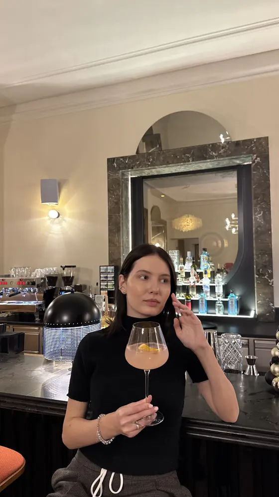 Me holding a stemmed glass with a pink drink at the hotel bar, with bottles and a mirror behind me.