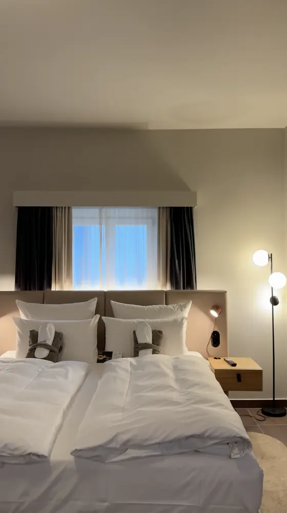 Bed with white bedding, a window behind the headboard and bedside lights in our Badeschloss junior suite.
