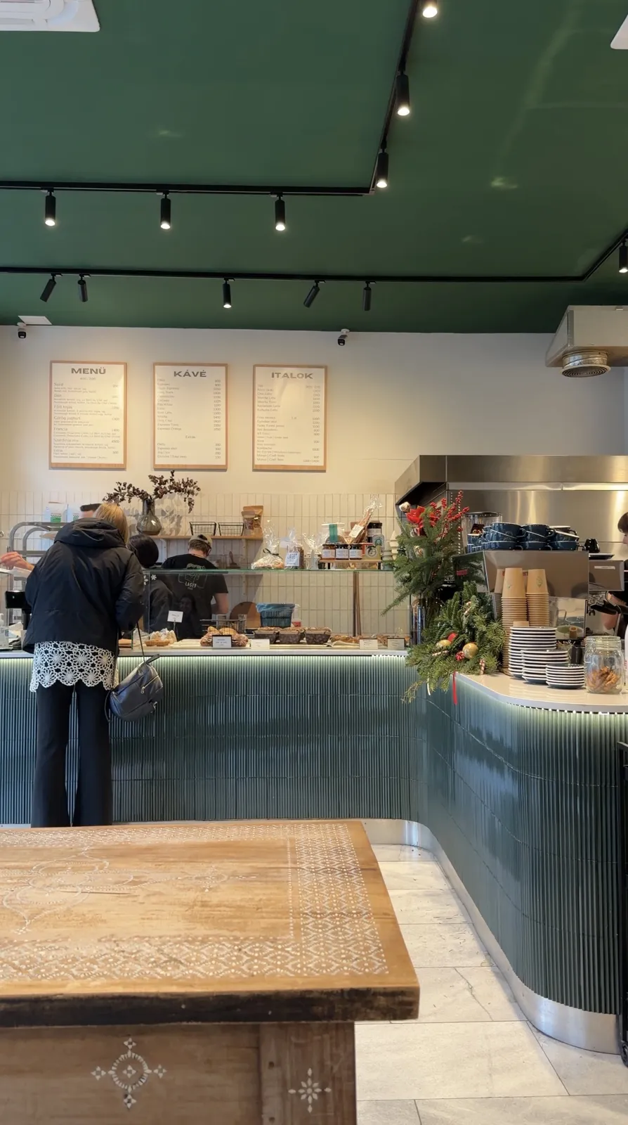 A café counter with a green front, menu boards on the wall and a baker behind it.
