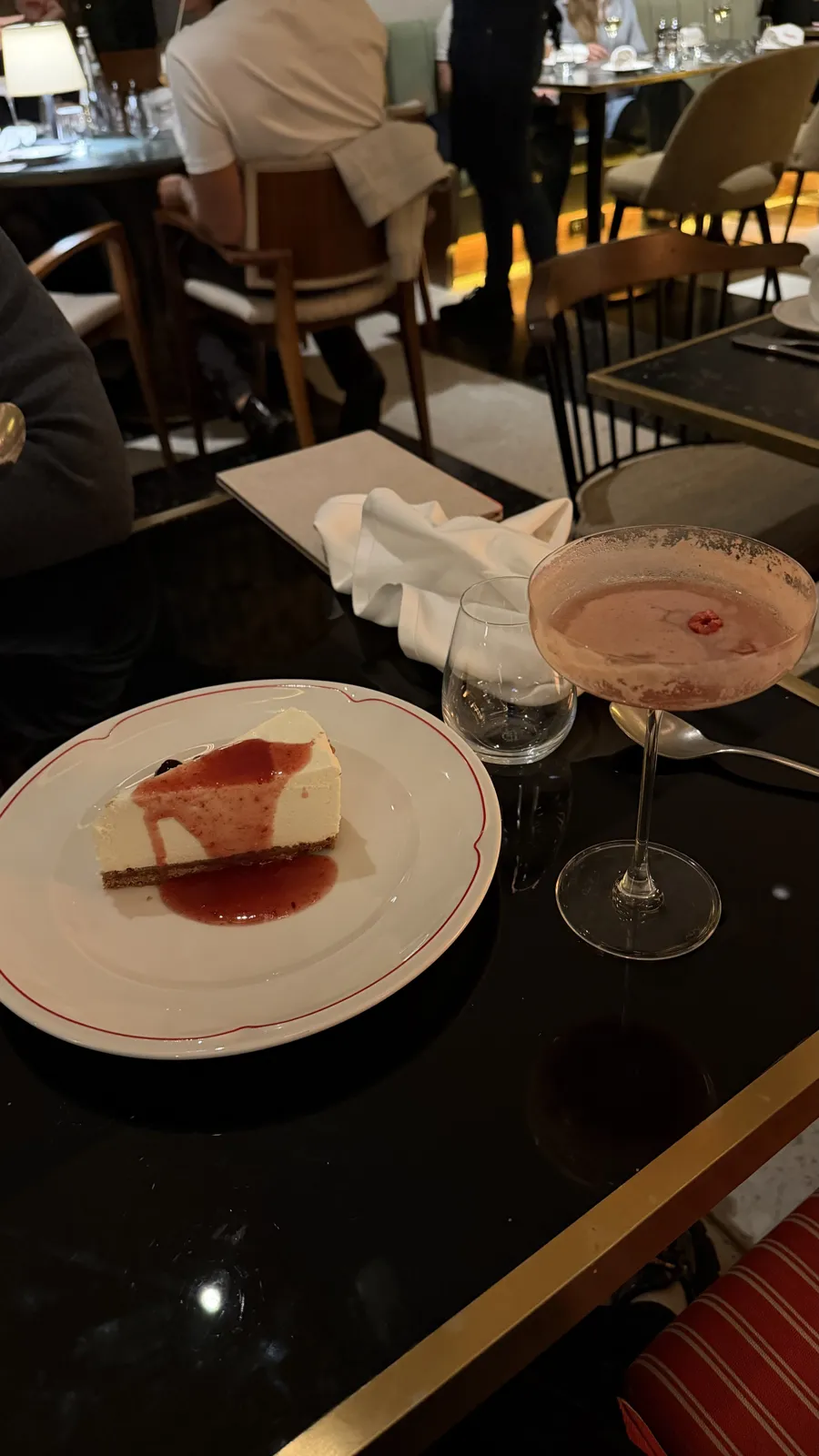 A slice of cheesecake with red sauce on a small plate beside a pink cocktail.