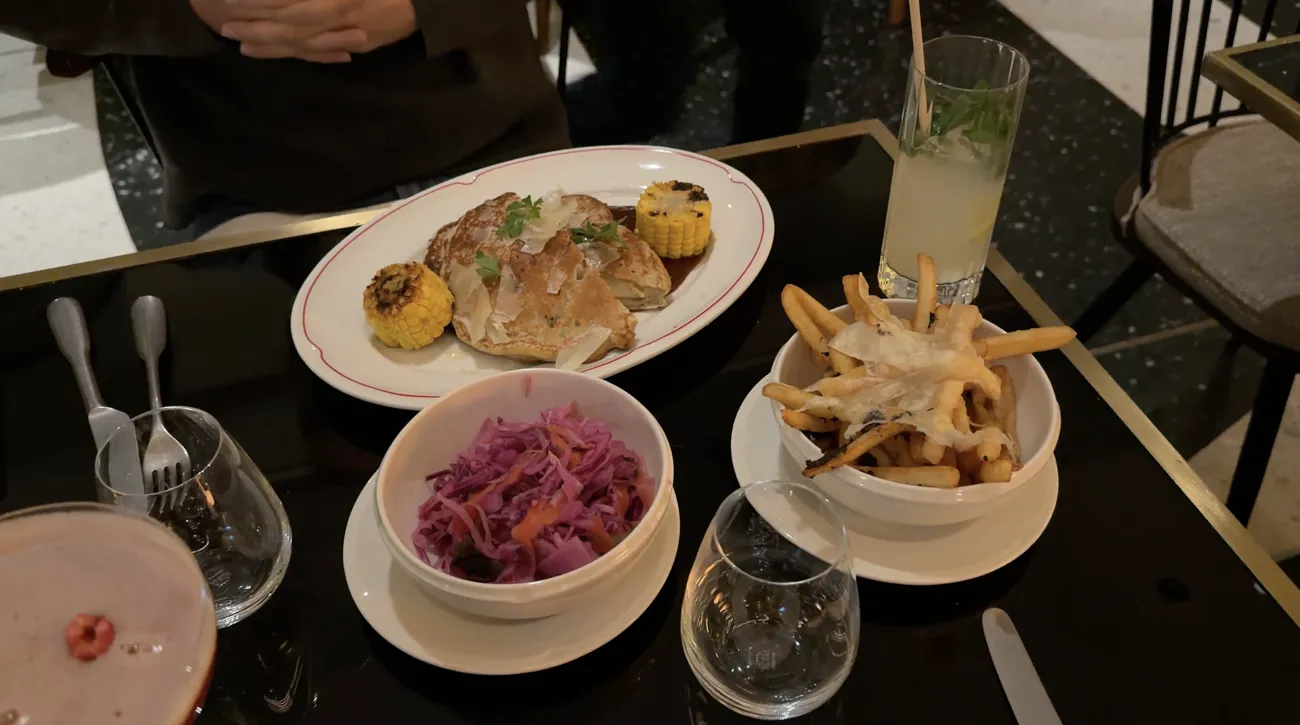 A plate of grilled chicken with corn, a bowl of red cabbage and a bowl of fries on a dark table.
