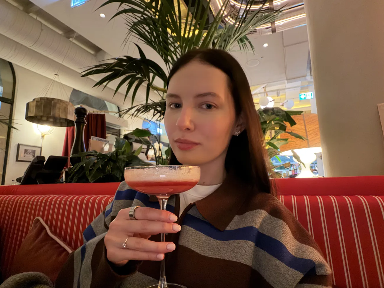 A woman holding a pink cocktail in a coupe glass, seated on a red striped banquette.