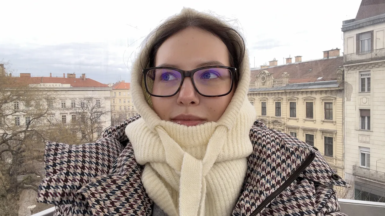 A woman in glasses, a cream hood and a checked coat with city buildings behind her.