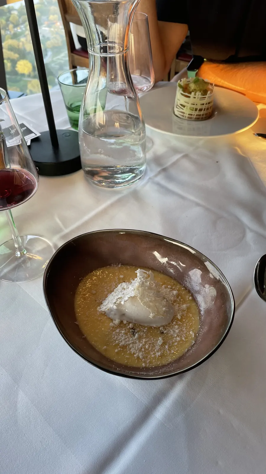 A yellow dessert with a white topping in a bowl, with another plated dessert across the table.
