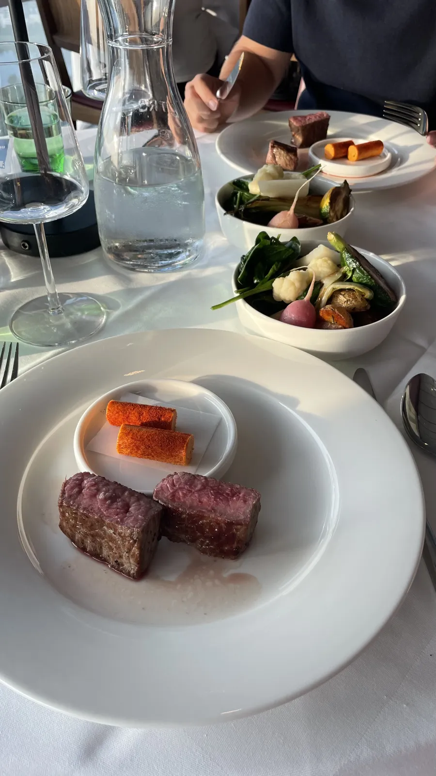 Two plates of pink-centred meat with separate bowls of vegetables on a white tablecloth.