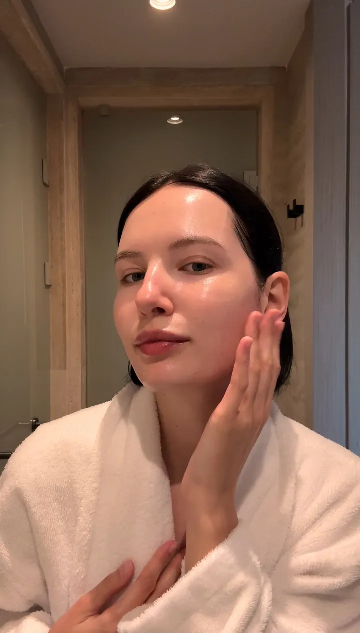 A woman in a white robe with a hand on her cheek, skin glowing after her skincare.