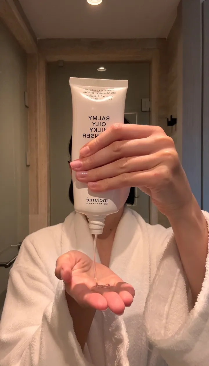 A hand holding a tube of Melumé Balmy Oily Milky Cleanser, squeezing some into the other palm.