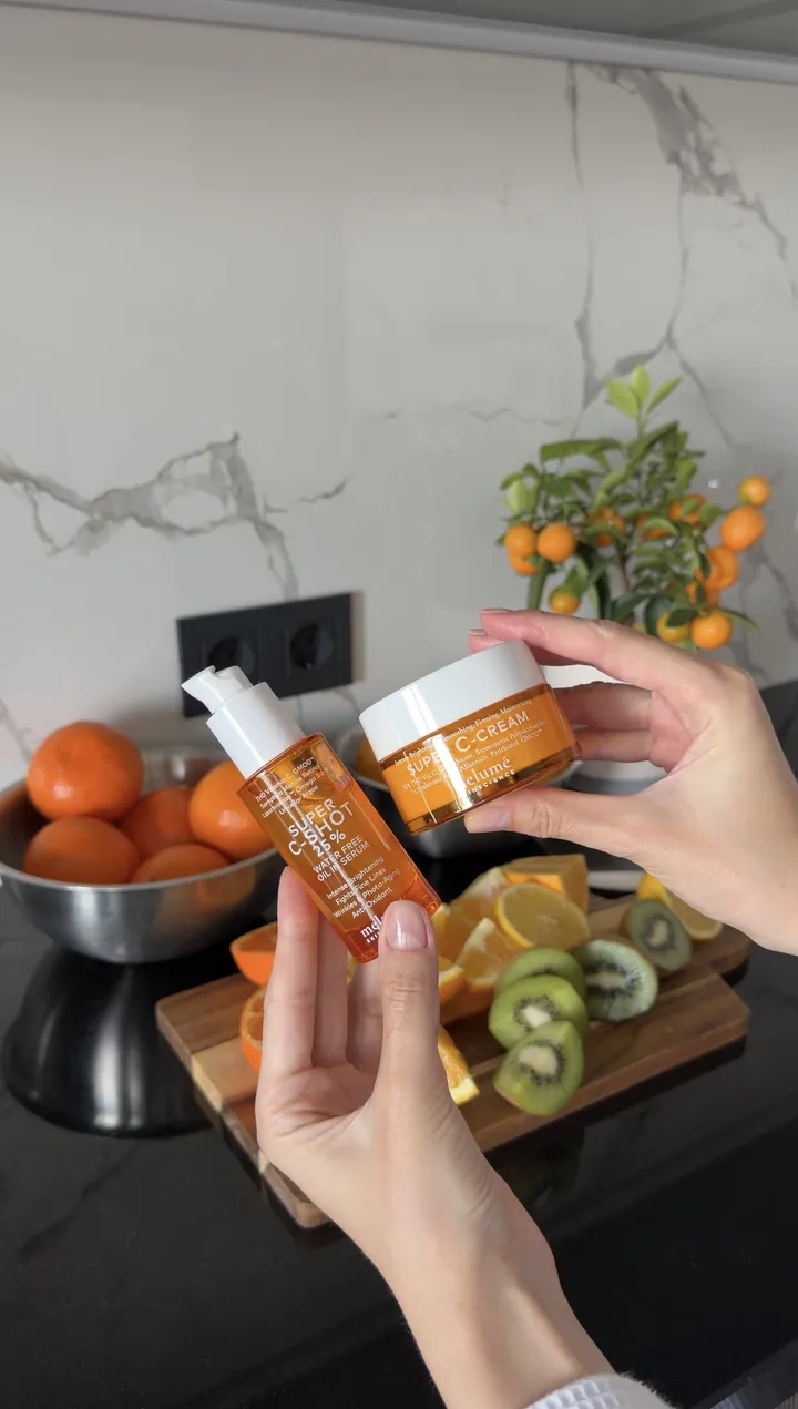 Hands holding an orange Melumé bottle and jar in front of a bowl of oranges.