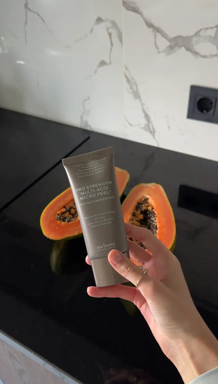 A hand holding a tube of Melumé Pro Strength Multi-Acid Micro Peel next to a halved papaya.