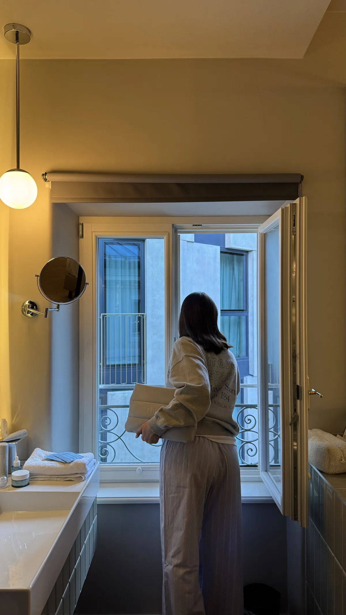 A woman in a sweatshirt and striped trousers stands at an open bathroom window, with a pendant light, a swing-arm mirror and towels by the basin.
