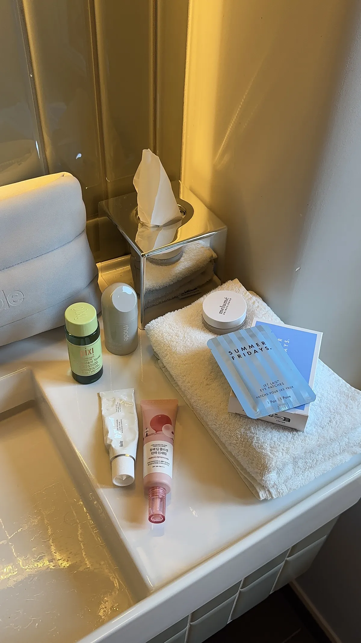 Skincare products on a bathroom shelf beside a basin, including a Pixi bottle, rhode and melumé products and Summer Fridays eye patches on a folded towel.