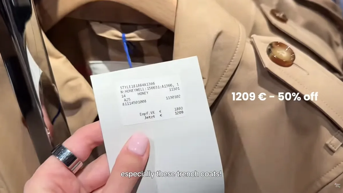 Me holding the label of a beige Burberry trench coat showing RRP €1,860 and €1,209, with an on-screen caption reading 1209 € - 50% off.