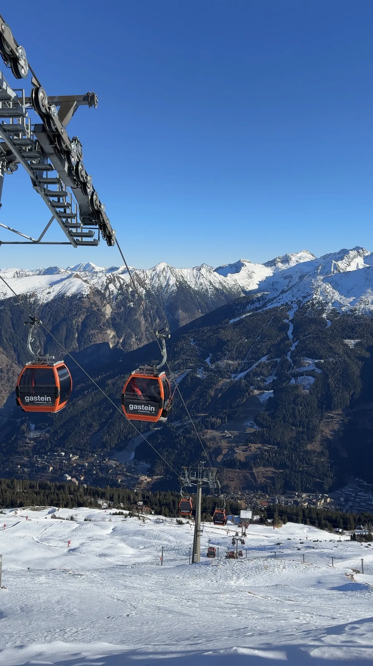 Red Gastein gondolas above a snowy slope, with a valley and snow-covered mountain ridges beyond.